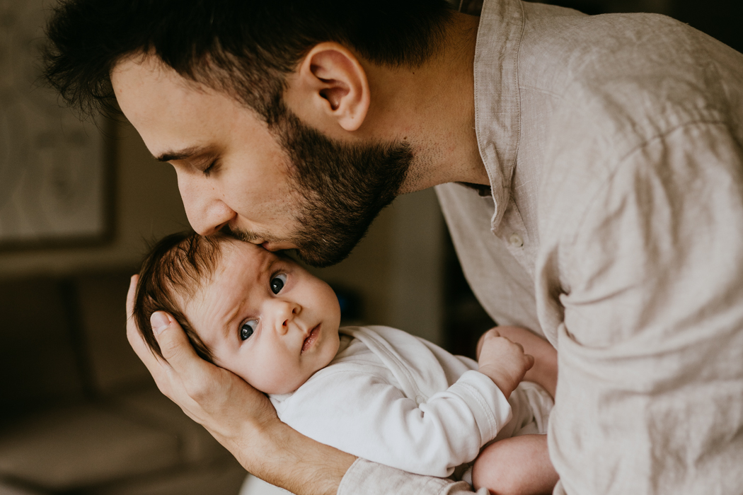 bliski kadr na którym młody tata trzyma na rękach noworodka i całuje podczas profesjonalnej sesji domowej